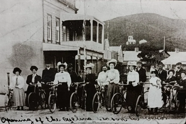 Black and white photograph of a group of women and bicycles standing outside a building in Coromandel town. Women are wearing full length black skirts, white tops and hats.